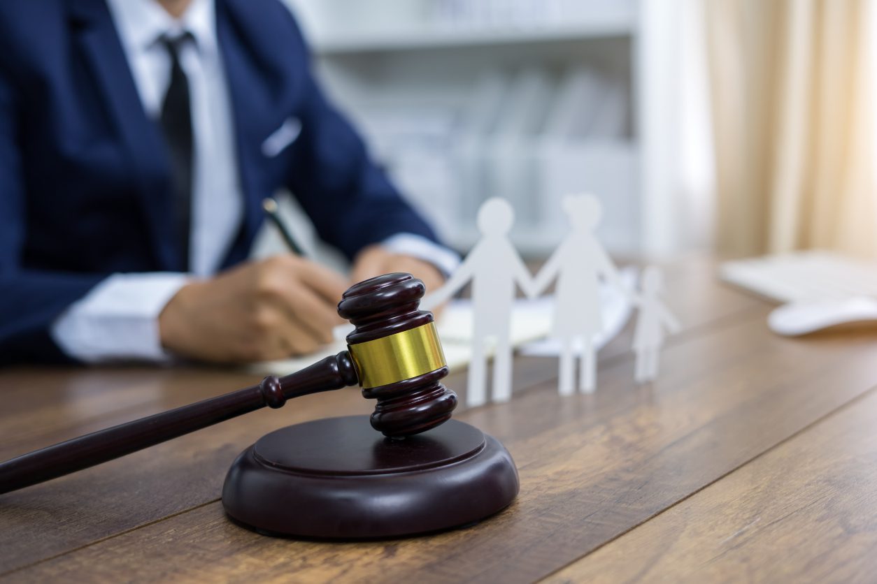 Gavel on desk with paper cutouts of a family in the background, and a person in a suit writing.