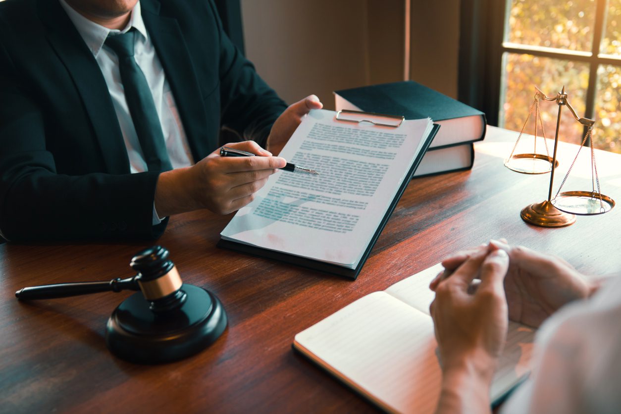 Two people discussing legal documents at a desk with a gavel, books, and scales of justice. One points with a pen, while the other holds their hands in front of a notebook.