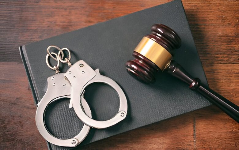 A gavel and handcuffs rest on a closed black book on a wooden surface.