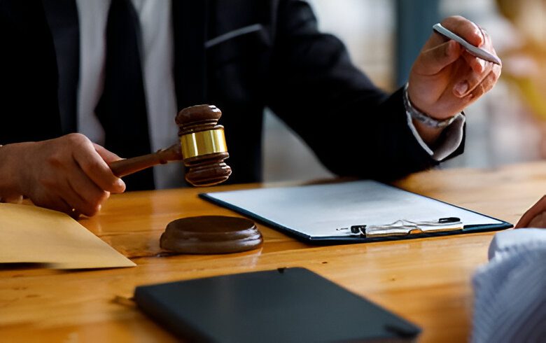 Person in a suit holding a gavel and reaching for a pen next to a clipboard and envelopes on a table.