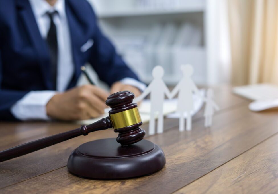 Gavel on desk with paper cutouts of a family in the background, and a person in a suit writing.